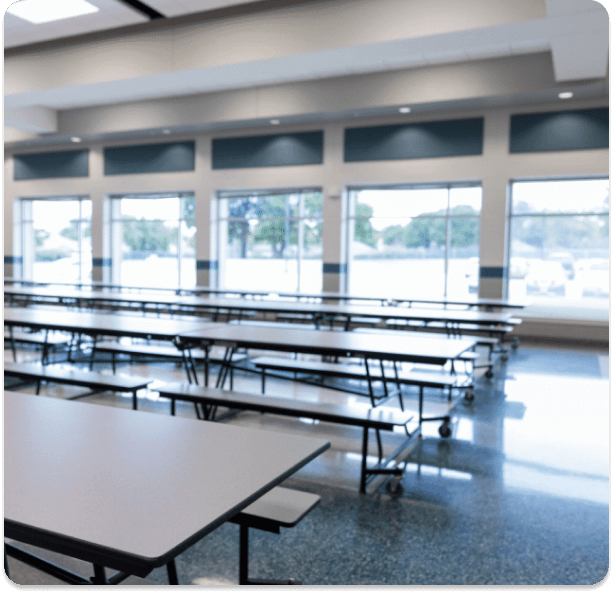 Empty school cafeteria with rows of folding tables beside a wall of windows